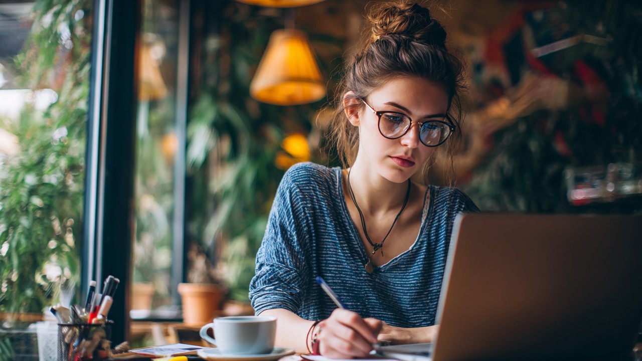 Content writer working on a laptop in a cozy cafe, surrounded by notes and coffee, researching hair transplant medical tourism.