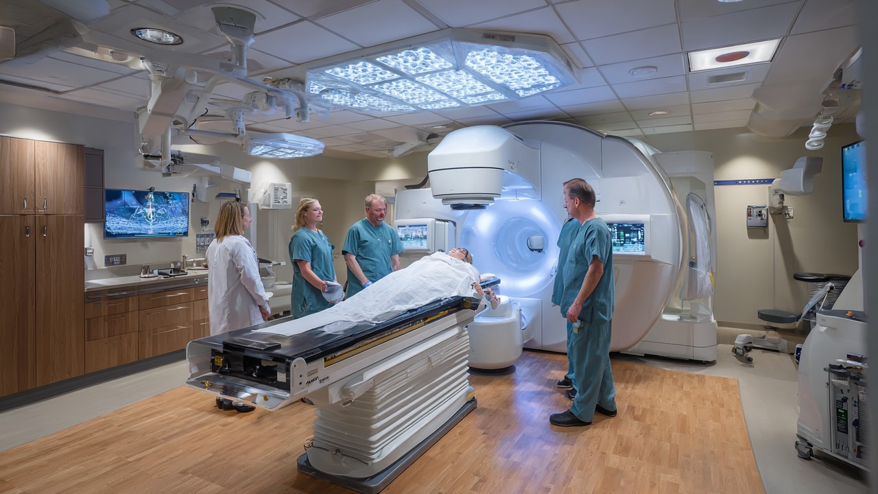 Pasukan perubatan di bilik rawatan onkologi berteknologi tinggi dengan mesin linear accelerator untuk terapi radiasi, dalam suasana klinik bersih dan terang.